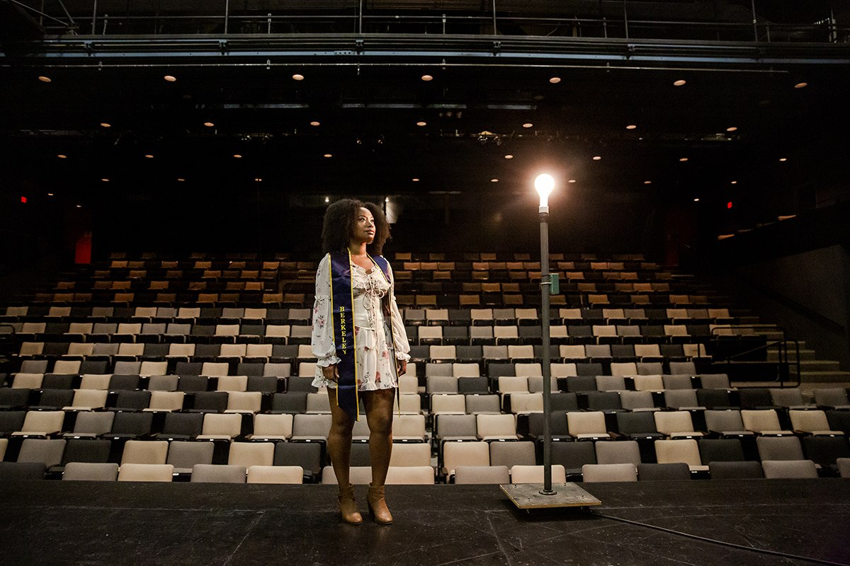 Anne Sharpe on an empty theater stage, wearing a white dress and blue Berkeley graduation stole.
