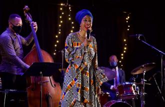 Jazzmeia Horn holds a mic and sings with a bassist and a drummer behind her. 