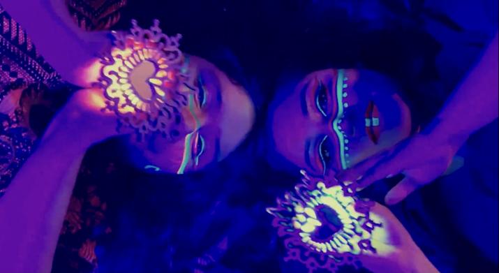Two women with glowing face paint lay on the ground looking at the camera holding masks.